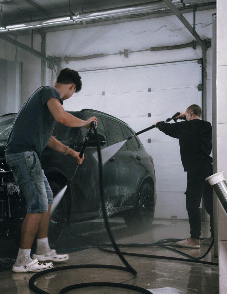 Two men using hoses to clean a black car in an indoor garage.